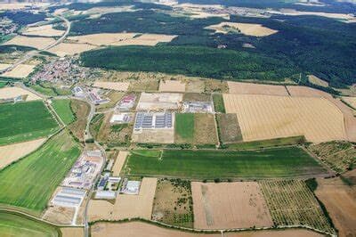 Weite Luftansicht von grünen Feldern, Industriegebäuden und einem Dorf vor einem bewaldeten Hintergrund.